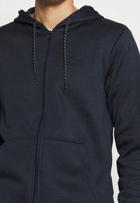 Marineblå hættetrøje med lynlås lavet af blødt materiale. Har en hætte med snore, to forlommer og en broderet logo på brystet.