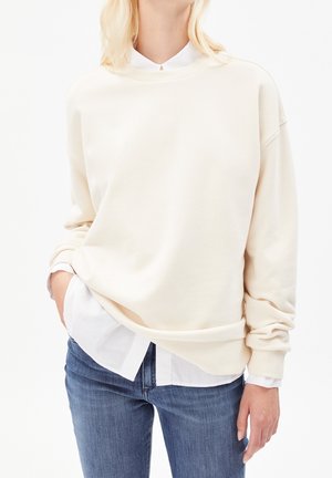 Frau trägt ein cremeweißes, übergroßes Sweatshirt über einem weißen Hemd mit Knöpfen und blauen Jeans, eine Hand in der Tasche.