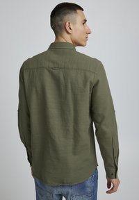 Camicia verde oliva con bottoni, maniche lunghe, tessuto testurizzato, dettaglio della spalla e polsini arrotolati con accenti di bottoni. Vestibilità casual.