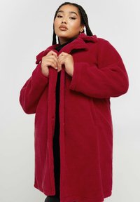Cappotto di pile rosso con colletto, tasche frontali e una texture morbida. Il cappotto ha una chiusura con bottoni e una vestibilità comoda.