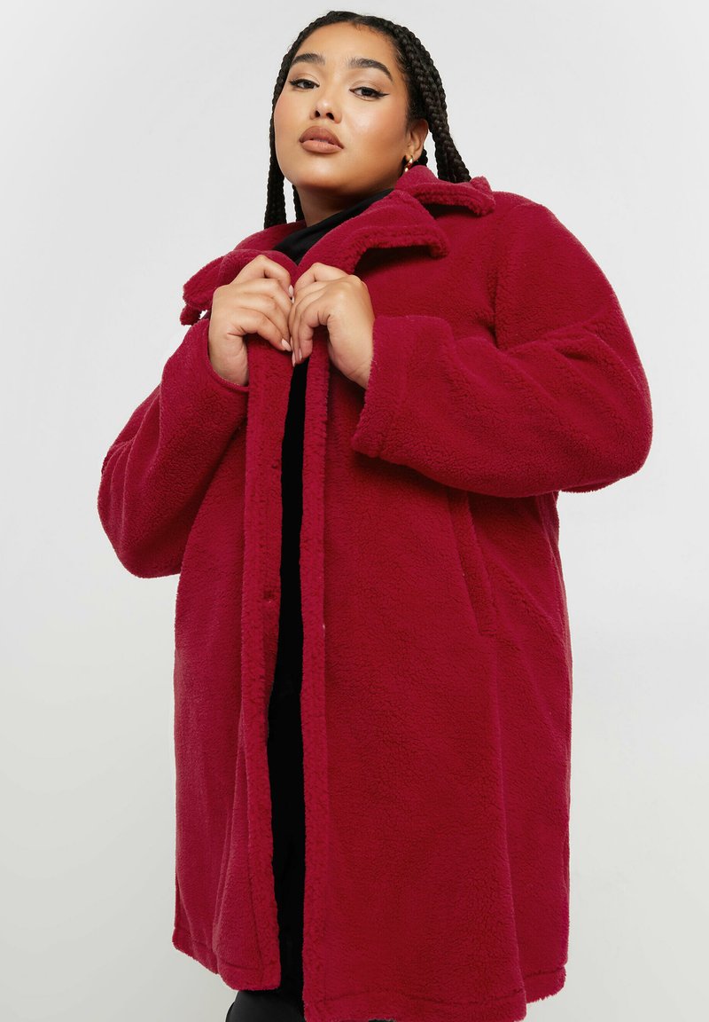 Cappotto di pile rosso con colletto, tasche frontali e una texture morbida. Il cappotto ha una chiusura con bottoni e una vestibilità comoda.