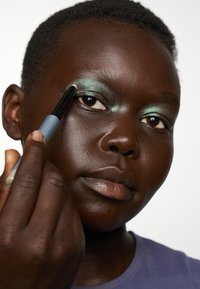 Gros plan sur une personne appliquant un fard à paupières vert scintillant avec un applicateur noir et bleu. La peau est d'un marron profond, les lèvres sont neutres. Fond texturé.