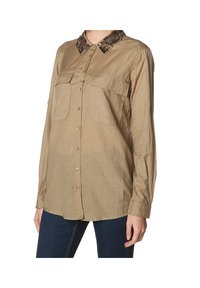 Camisa de manga larga beige clara con un cuello marrón estampado, dos bolsillos frontales y cierre de botones; la tela parece ligera y ligeramente texturizada.