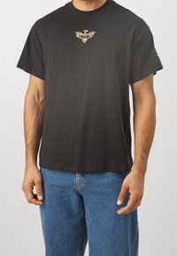 Czarna bawełniana koszulka z krótkim rękawem, z złotym emblematem na klatce piersiowej oraz luźnym krojem. Połączona z niebieskimi dżinsami.