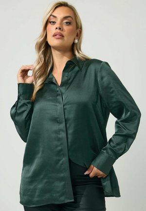 Vrouw met lang blond haar, die pareloorbellen en een donkergroene satijnen blouse met lange mouwen draagt tegen een effen achtergrond.