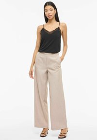 Czarna kamizelka z koronkową lamówką, w zestawie z beżowymi szerokimi spodniami. Modelka ma na sobie czarne sandały na obcasie z złotym akcentem.