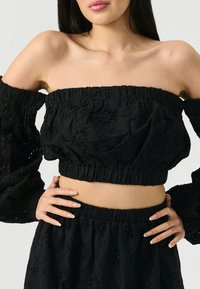 Čierny crop top bez ramienok a zodpovedajúca sukňa, ktorá má textúrovanú látku s kvetinovou výšivkou a smockovanými detailmi.