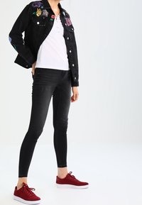 Chaqueta de mezclilla negra con bordado floral, camisa blanca con cordones, jeans skinny negros y zapatillas rojas con suelas blancas.