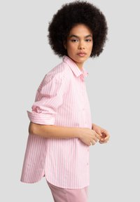 Camisa de botones de color rosa claro con rayas verticales blancas, mangas enrolladas y dobladillo curvado. Tejido de algodón texturizado.
