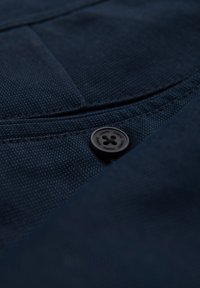 Tessuto blu navy con un motivo testurizzato, caratterizzato da un bottone nero rotondo con branding inciso. Comprende cuciture visibili lungo i bordi.
