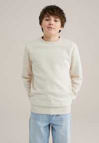 Svetlo bež pulover s krojem okoli vratu, z ribastimi manšetami in robom. Ima gladko teksturo in zategnjeno obliko, v kombinaciji s svetlo modrimi džins hlačami.