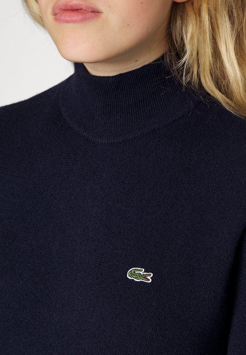 Pull à col roulé marine en tissu doux, avec un petit logo de crocodile brodé en vert et blanc sur la zone de la poitrine gauche.