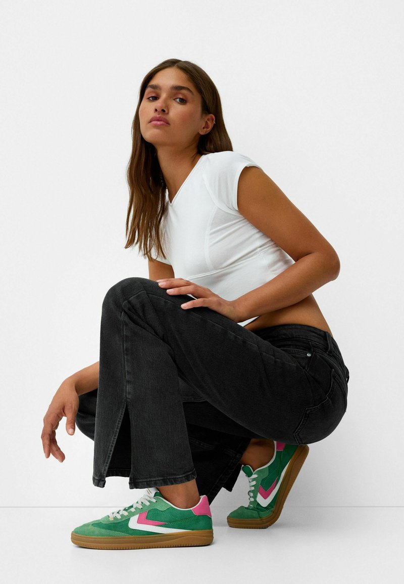 Giovane donna accovacciata che indossa una T-shirt bianca aderente, jeans neri con fenditure anteriori e sneaker verdi con accenti rosa e bianchi.