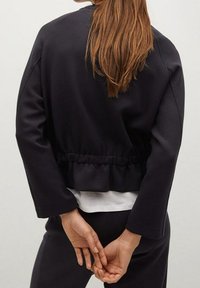 Sudadera negra, corta, con una cinturilla fruncida, mangas largas y textura suave. Llevada sobre una parte superior blanca, vista desde la parte de atrás.