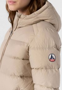 Frau, die eine beige Steppjacke mit Kapuze trägt, mit sichtbarem JOTT-Logo-Patch am Ärmeln und einem Frontreißverschluss.