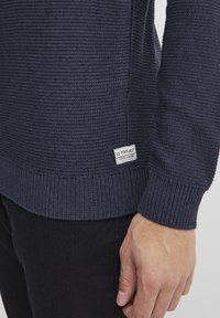 Maglione blu navy in maglia con tessuto testurizzato, polsini e orlo a coste. Etichetta sul lato con la scritta "11 PROJECT." Pantaloni neri parzialmente visibili.