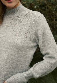Pelēks adīts džemperis ar augstu kakla daļu, vienā plecā rotāts ar dekoratīvām sudraba pērlītēm, veidojot teksturētu dizainu.