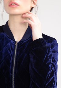 Giubbotto bomber in velluto trapuntato blu navy con polsini e colletto a costine, dotato di una zip centrale. Tessuto liscio e motivo geometrico visibile.
