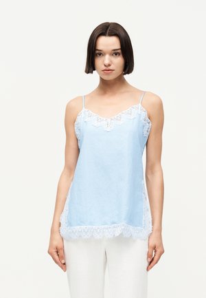 Νέα γυναίκα με κοντά σκούρα μαλλιά που φοράει ανοιχτό μπλε αμάνικο τοπ με λευκή δαντέλα και άσπρο παντελόνι, μπροστά σε μονόχρωμο φόντο.