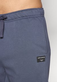 Tamsiai mėlynos sportinės kelnės, pagamintos iš minkštos medžiagos, su raišteliu juosmenyje ir juodu prekės ženklo etikete šone.