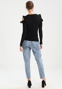 Zwart geribd topje met cold shoulder uitsparingen en ruchedetails; gecombineerd met lichtblauwe rechte spijkerbroek en zwarte hoge hakken.