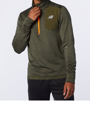 Mann trägt ein olivgrünes langärmeliges Sportoberteil mit orangefarbenem Viertelreißverschluss und Brusttasche, kombiniert mit schwarzen Hosen.
