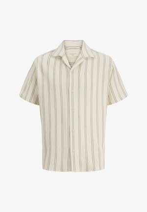 Adolescente che indossa una camicia a maniche corte crema a righe con bottoni e pantaloncini abbinati, calzini bianchi, sneaker e una bandana rossa legata intorno al collo.