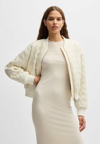 Quilted off-white bomber jacket with ribbed cuffs, high collar, and front zipper. Worn over a fitted beige dress, highlighting texture contrast.