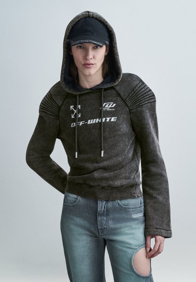 Άτομο που φοράει γκρι σκούρο φούτερ Off-White με ανεβασμένο το καπό, μαύρο καπέλο και μπλε τζιν με μεγάλη κυκλική τρύπα σε ένα μηρό.