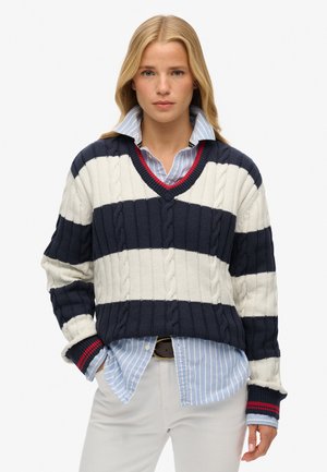 Marineblauwe en witte gestreepte gebreide trui met een textuur van kabelontwerp, V-hals, en rode accenten op de manchetten over een blauw gestreepte overhemd met kraag.