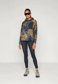 Hettegensere i blå og brun tie-dye, med en lomme foran; kombinert med svarte leggings og grå joggesko.