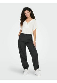 Casual outfit bestaande uit een lichtcrème blouse met een ritssluiting in de hals en zwarte cargobroeken met zijzakken. Witte sneakers maken de look af.