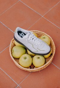 Baskets blanches posées sur des pommes vertes à l'intérieur d'un panier tressé sur un sol carrelé.