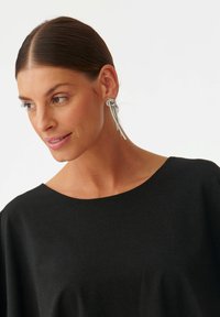 Srebrni uhani z obliko vozla, ki vključujejo dva podolgovata trakova. Model nosi črno, majico s kratkimi rokavi.