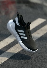 Scarpa sportiva in bianco e nero con tre strisce bianche, sospesa sopra l'asfalto con segnaletica stradale bianca e detriti neri sparsi.