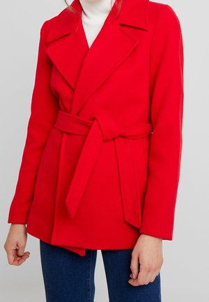 Femme portant un manteau rouge vif ceinturé sur un pull à col roulé blanc et un jean bleu, debout devant un fond clair uni.