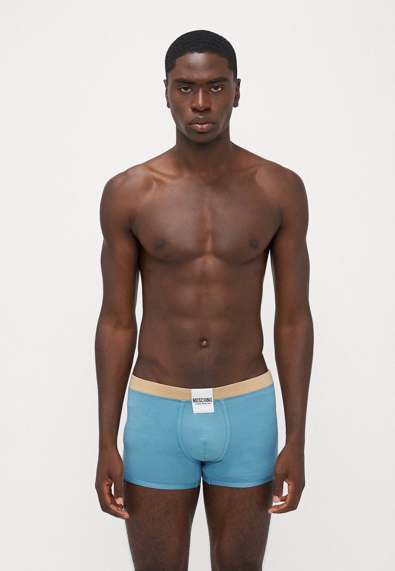 Blue cotton boxer briefs with a beige waistband, featuring a Moschino logo tag at the front. The fit is snug and the fabric has a smooth texture.