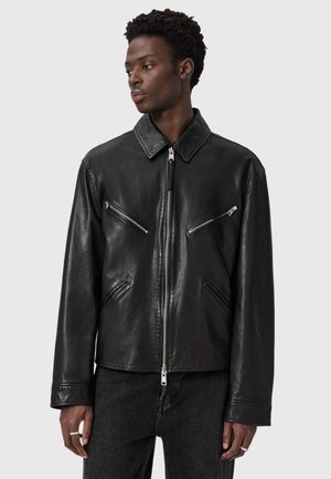 Junger Mann trägt eine schwarze Lederjacke mit Reißverschluss und schrägen, mit Reißverschluss versehenen Brusttaschen, steht vor einem einfarbigen hellgrauen Hintergrund.