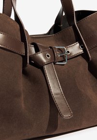 Primer plano de un bolso de mano de ante marrón con correas de cuero marrón oscuro y una hebilla plateada.