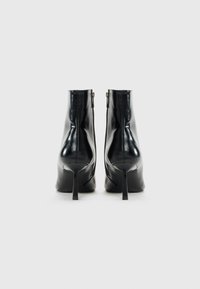 Bottines en cuir verni noir avec des talons aiguilles pointus et des zips latéraux, vues de l'arrière sur un fond blanc.