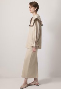 Beige top s volánkovým límcem a nabíranými rukávy, zkombinovaný s odpovídající dlouhou sukní. Modelka má na sobě sandály na podpatku s otevřenou špičkou a výřezy.