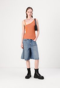Oranje geribbelde tanktop met asymmetrische halslijn, gecombineerd met lichtblauwe denimshorts. Zwarte veterschoenen met een robuuste uitstraling en een kleine zwarte tas.