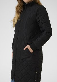 Manteau long matelassé noir avec un col haut, une fermeture éclair à l'avant et des poignets élastiques ; comporte deux poches latérales et des accents de boutons-pression.