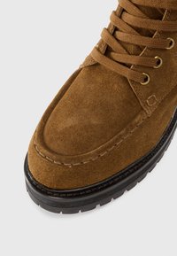 Boot din suedă maro, cu șireturi, vârf rotund, talpă groasă din cauciuc negru și detalii cusute în partea superioară a pantofului.