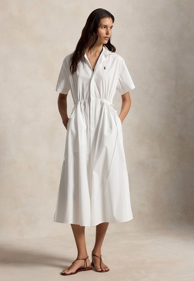 Femme portant une robe mi-longue blanche à manches courtes avec col, taille cintrée, poches et sandales plates marron, debout devant un fond neutre.