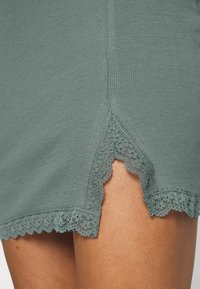 Vêtement en tissu côtelé gris avec un ourlet décoratif en dentelle, présentant des bords festonnés qui laissent entrevoir la peau en dessous. Texture douce avec une coupe ajustée.