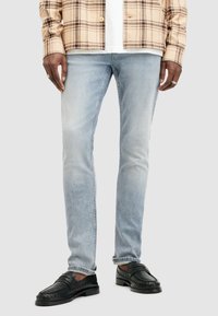 Jeans de mezclilla azul claro con un corte slim y textura desgastada, combinados con mocasines de cuero negros. El modelo lleva una camisa de cuadros beige.
