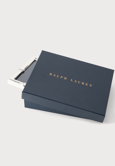Κουτί ναυτικού μπλε με χρυσό κείμενο "Ralph Lauren", μερικώς ανοιχτό, που αποκαλύπτει μέσα μια ασημένια μεταλλική αγκράφα ζώνης.
