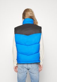 Person med långt hår som bär en blå och svart pufferväst över en beige långärmad tröja och ljusblå jeans, vänd mot en vit bakgrund.