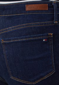 Tamsiai skalbti džinsai su rudu odiniu žymėjimu, kuriame užrašyta "TOMMY HILFIGER". Matomi oranžiniai siūlai ir mažas logotipas ant užpakalinio kišenės.